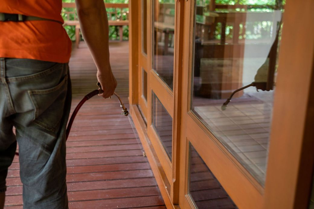 A person in an orange shirt and blue jeans uses a spray nozzle to apply treatment along the base of a wooden door frame.
