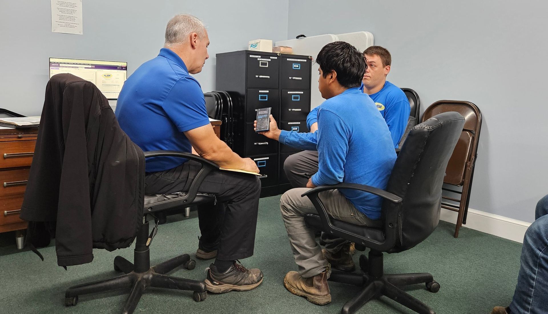 An adult in a blue shirt discusses something with two youths in blue shirts while sitting in an office with file cabinets.