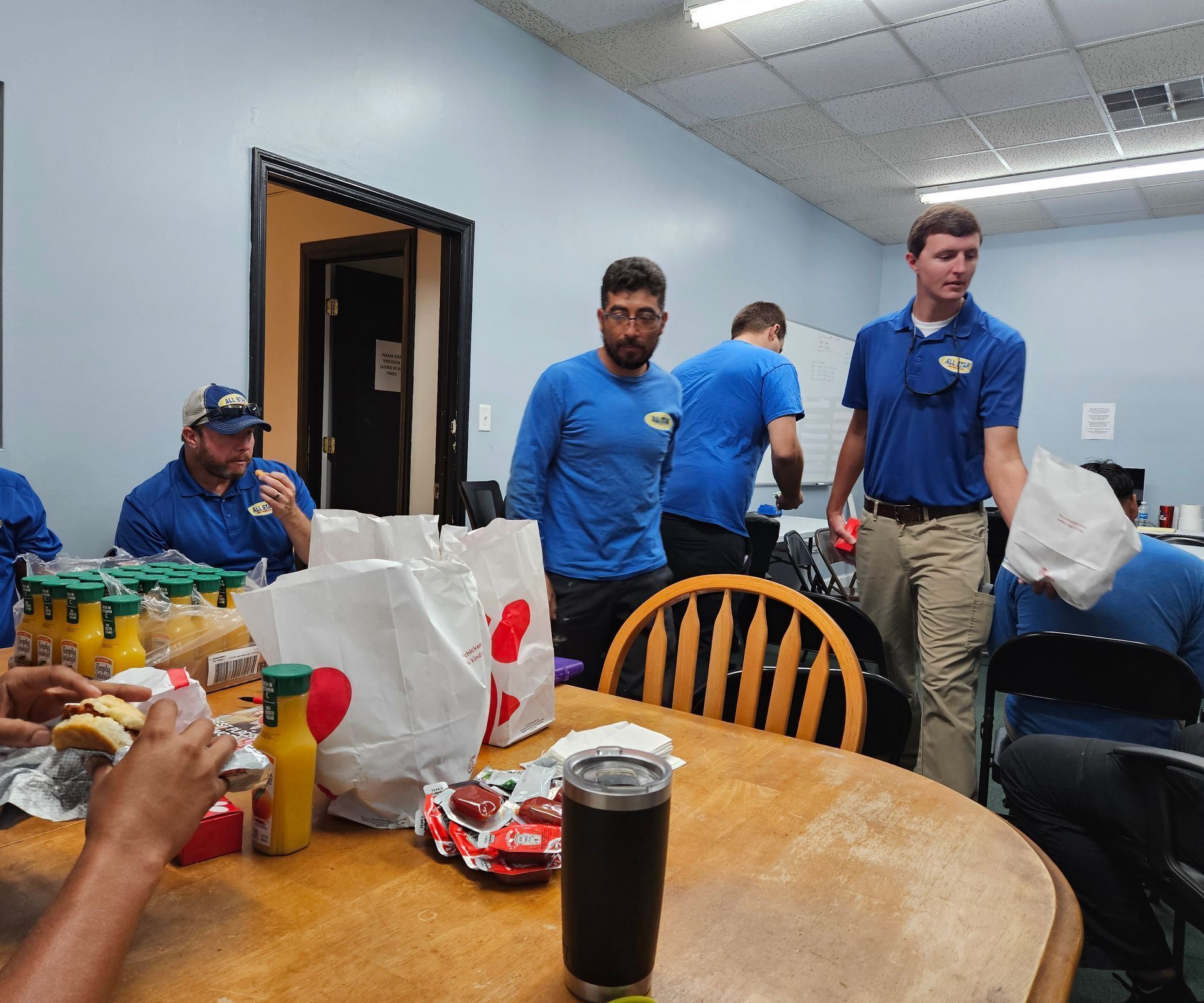 A group of people wearing blue uniforms distribute Chick-fil-A takeout bags at a wooden table in an office setting.