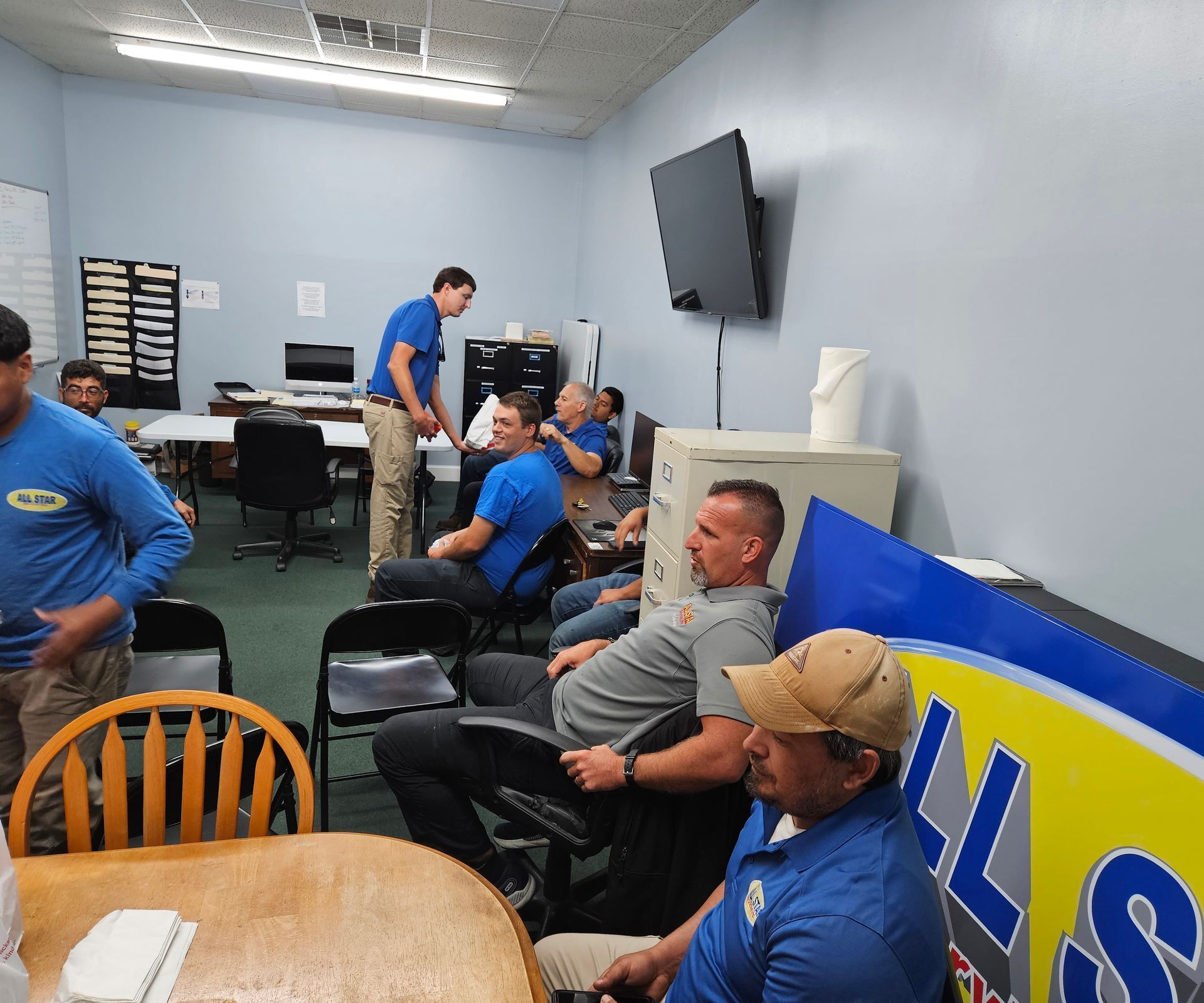 A group of people in a casual office setting meeting around tables, with some seated and one standing.