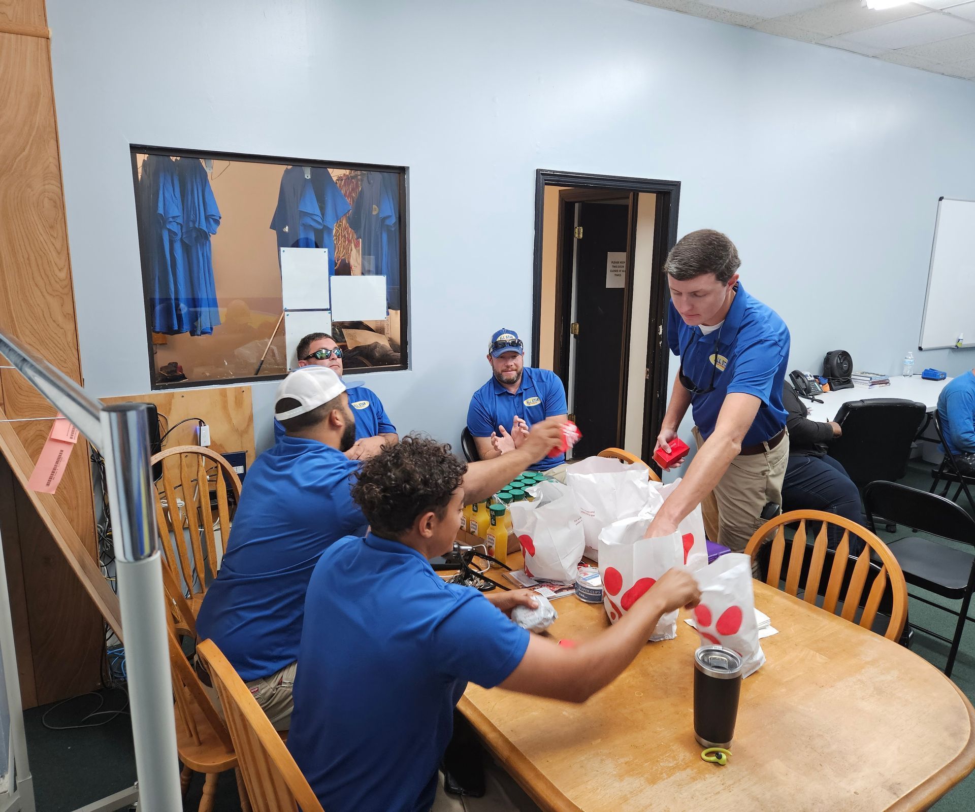 A group of people wearing blue shirts sit around a wooden table in an office, packing Chick-fil-A bags.