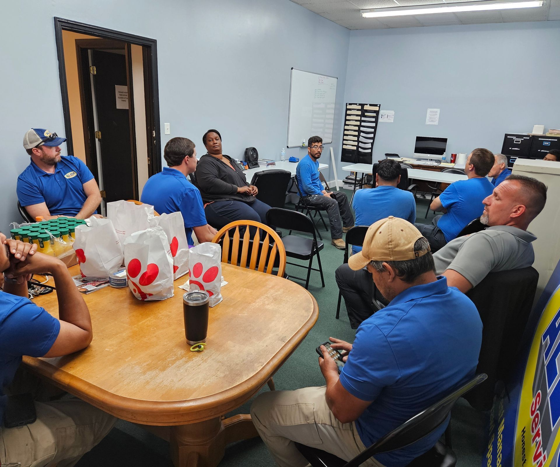 A group of people in a breakroom sit around a wooden table with Chick-fil-A bags during a meeting.