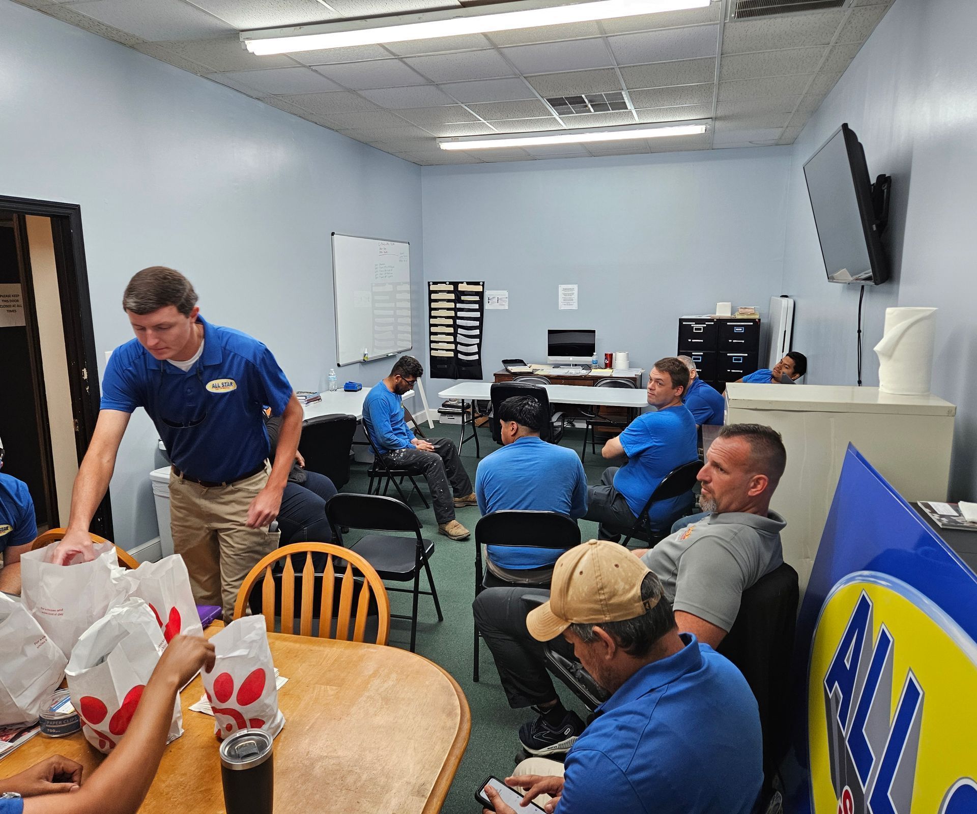 A group of people wearing blue shirts sit in a breakroom, some eating Chick-fil-A while others listen to a presentation.