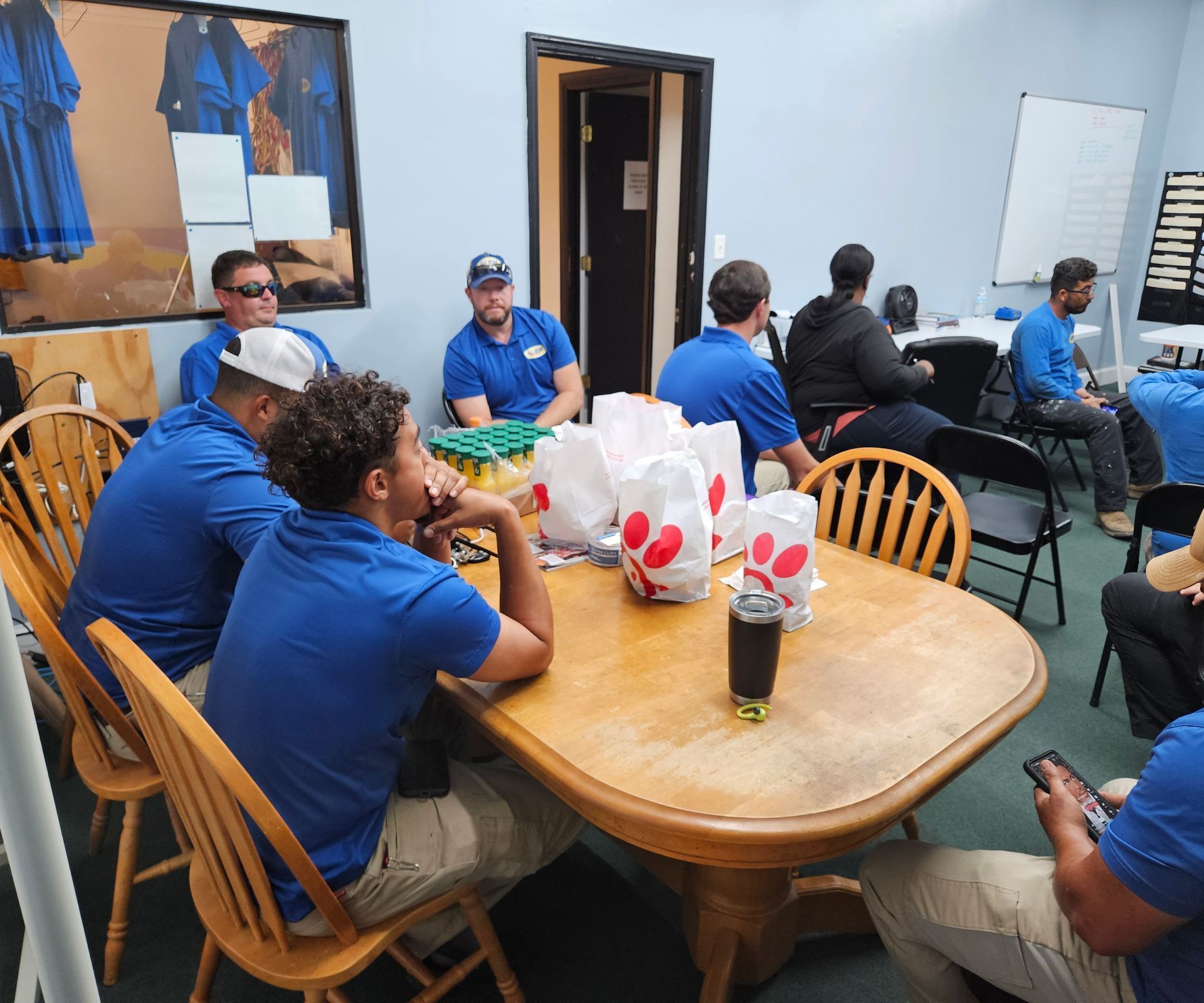 A group of people wearing blue shirts sit around a wooden table in an office, eating lunch from Chick-fil-A bags.