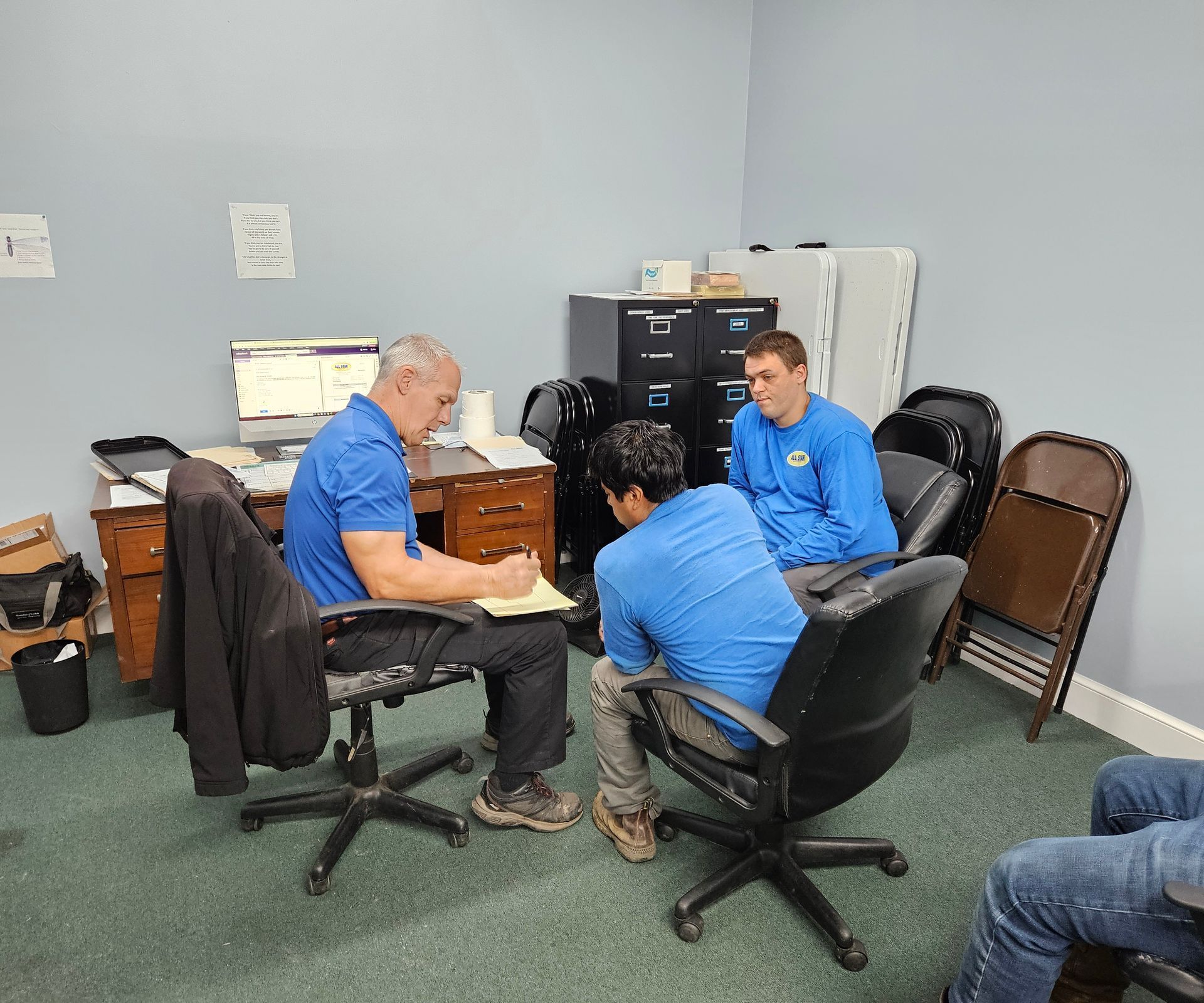 Three people in blue shirts have a meeting in an office, with one person writing at a desk and two seated in chairs.