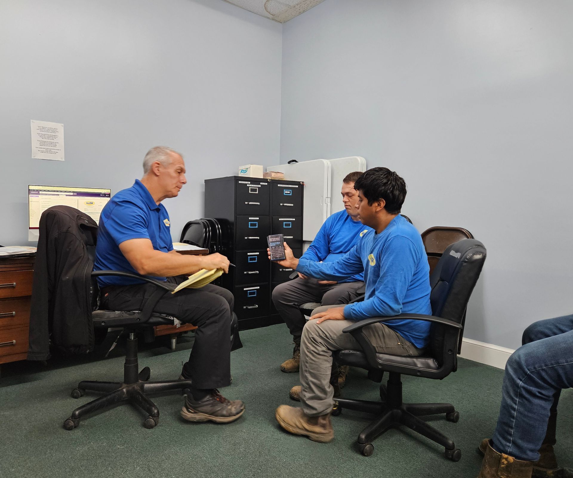 Three people in blue shirts have a professional discussion while sitting in an office with filing cabinets.