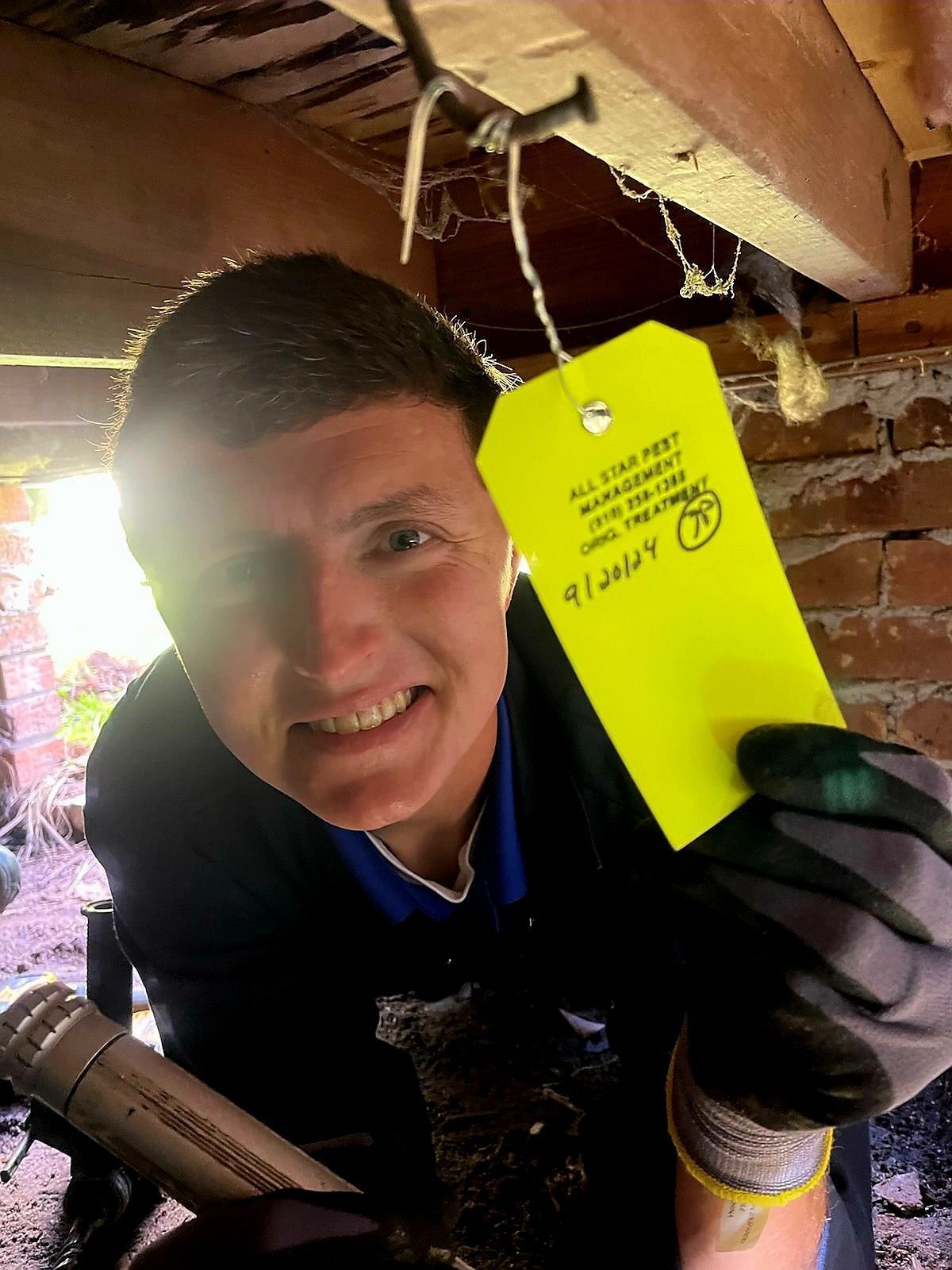 A person in a crawl space holds a yellow pest control tag hanging from a floor joist, smiling at the camera.