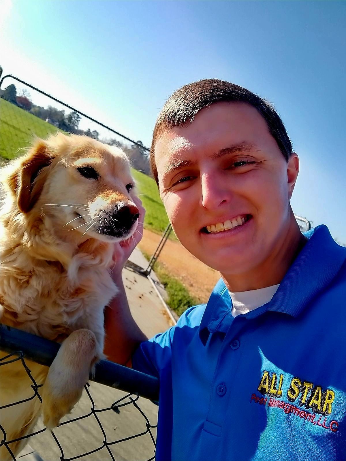A smiling person in a blue polo shirt poses for a selfie next to a golden dog resting its paw on a chain-link fence.