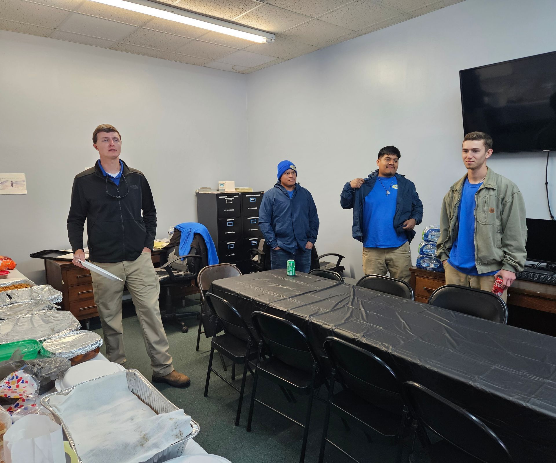 Four people stand in an office breakroom, positioned near a long table with catering containers and a television.