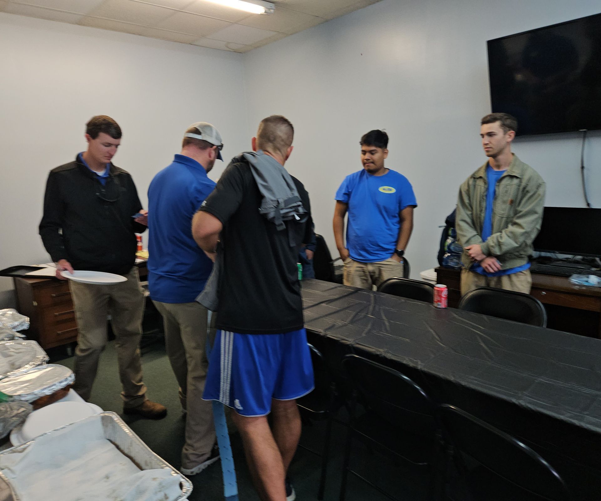 Five people stand around a black table in a plain, indoor room, some looking at papers while others stand by the table.