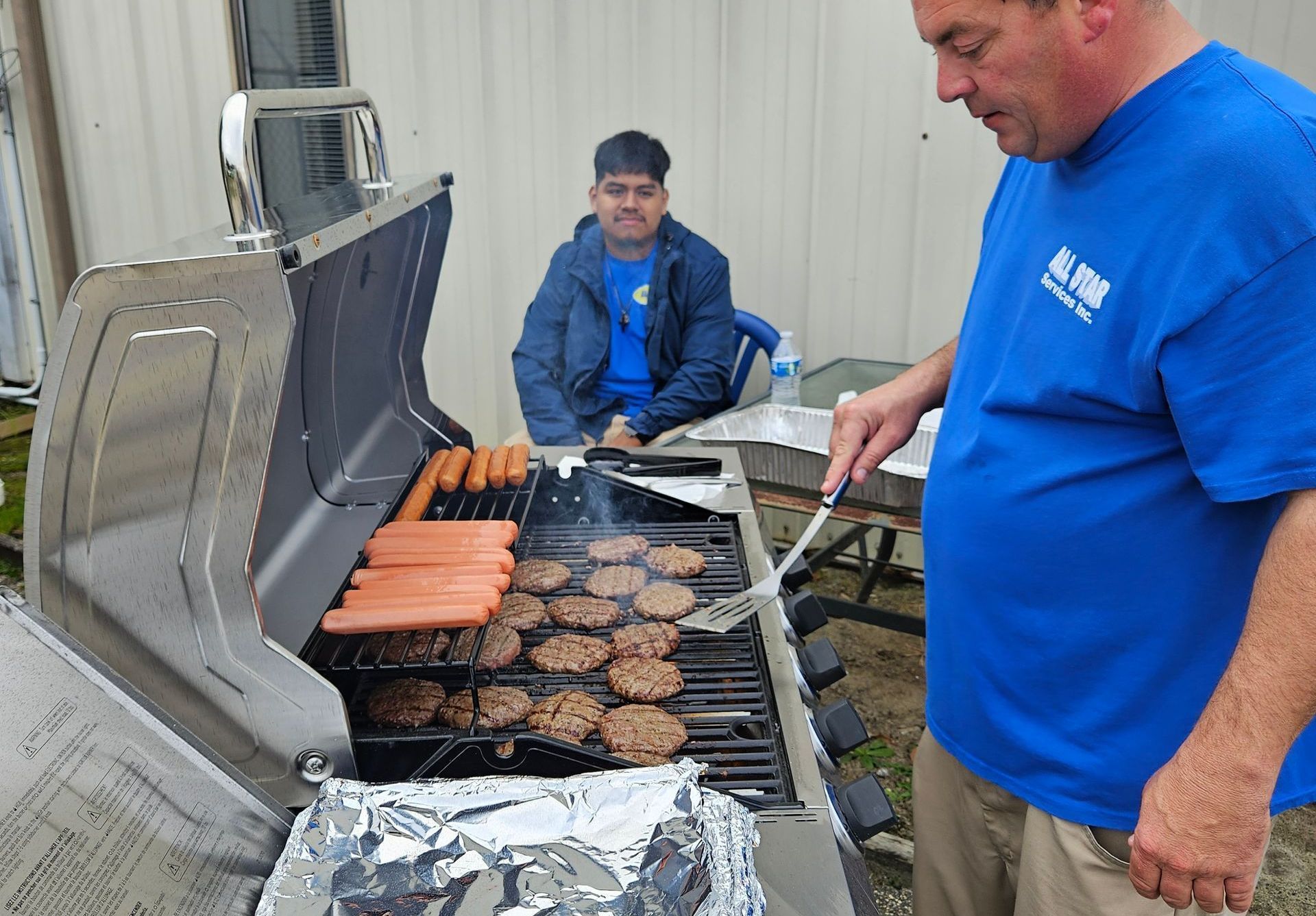 A person in a blue t-shirt grills hot dogs and hamburger patties while another person sits nearby outdoors.