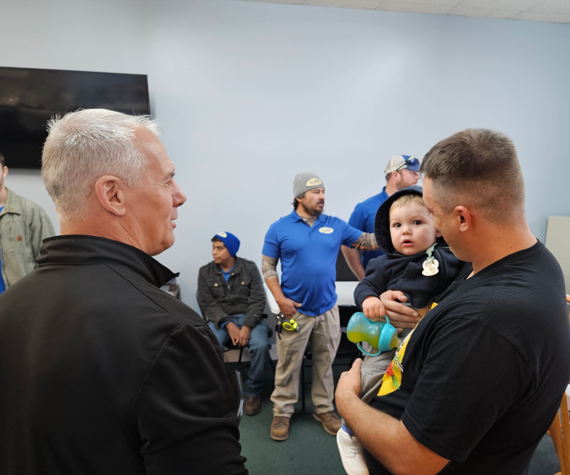 A group of people standing in an indoor setting, with one person holding a small child in a hoodie and holding a bottle.