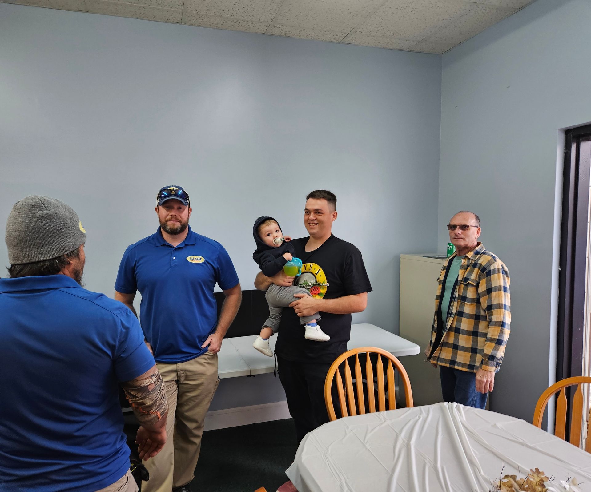 Four people, one holding a toddler, stand in a room with light blue walls and folding tables.