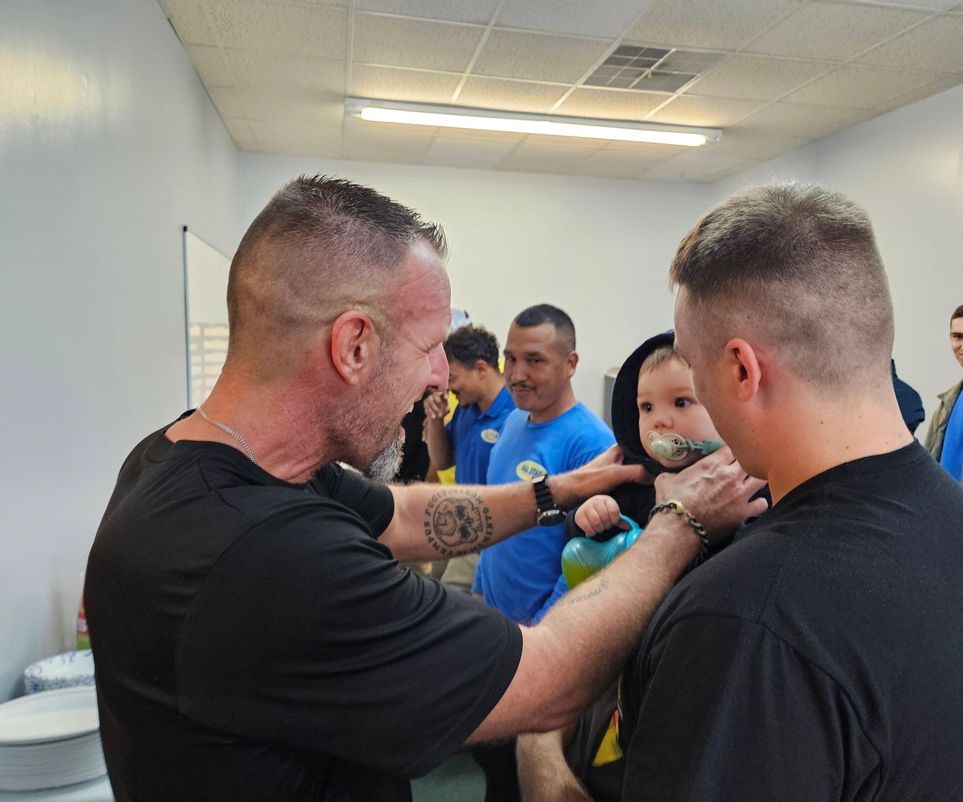 Two men in black shirts interacting with a small child in a black hooded jacket inside a brightly lit room.