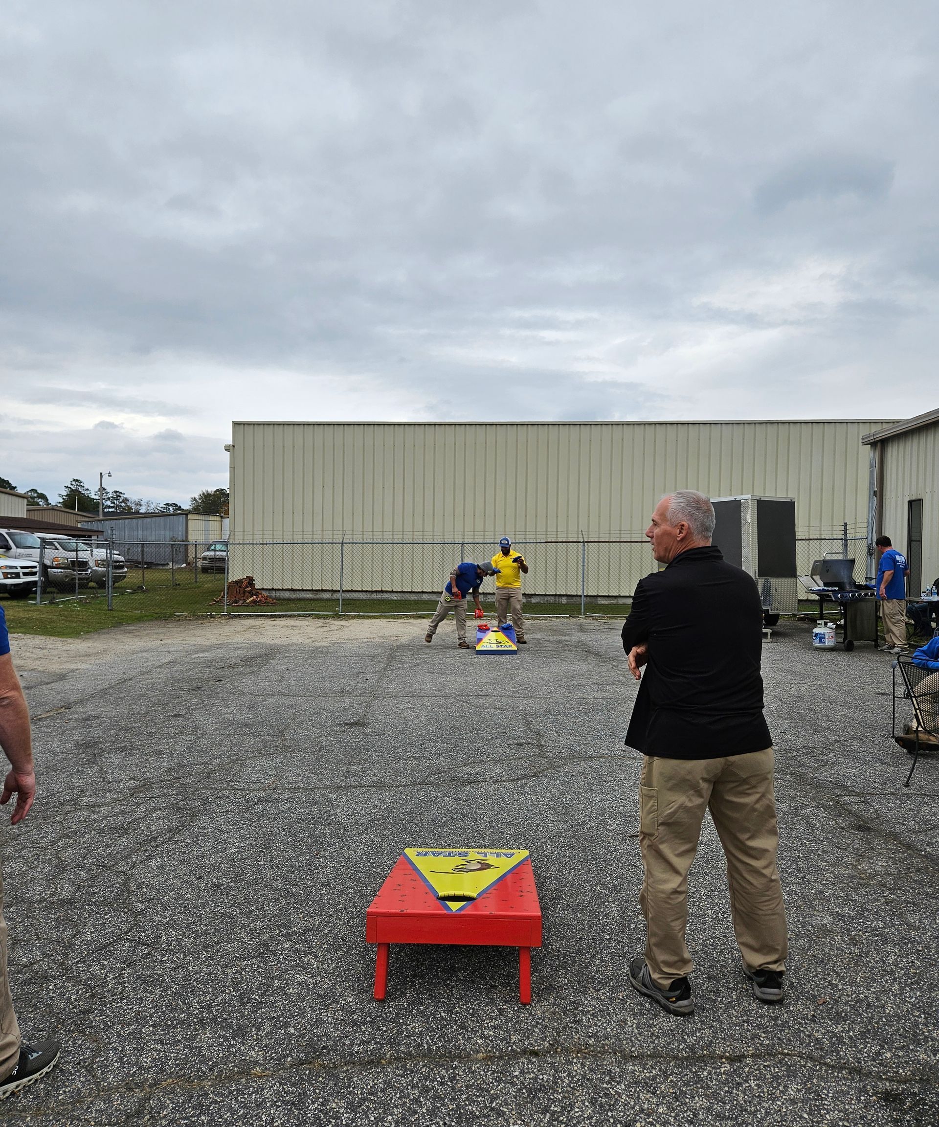 People playing cornhole on a gravel lot outside a large warehouse on an overcast day.