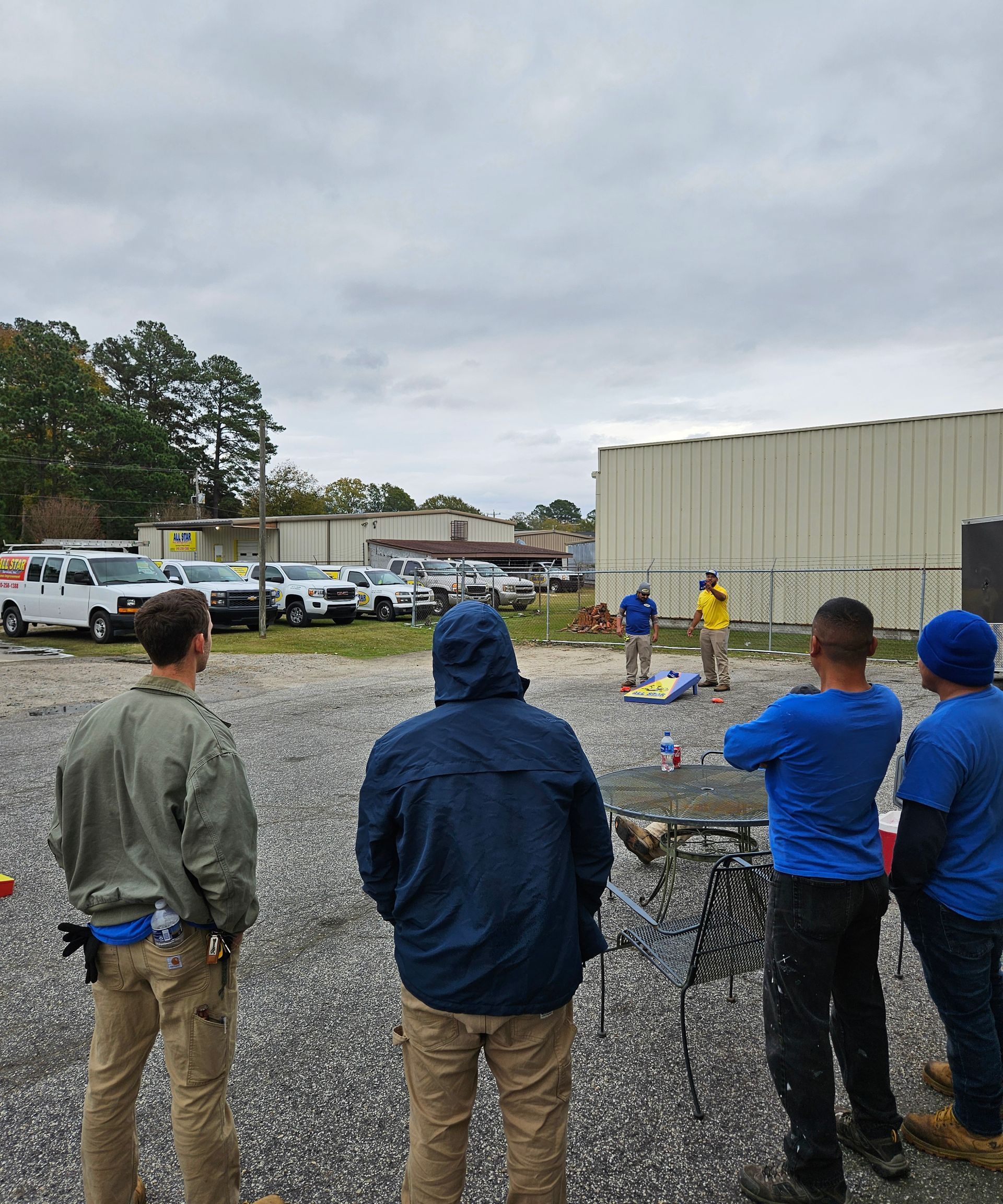 A group of people standing on a gravel lot outdoors, watching two individuals play a game of cornhole.
