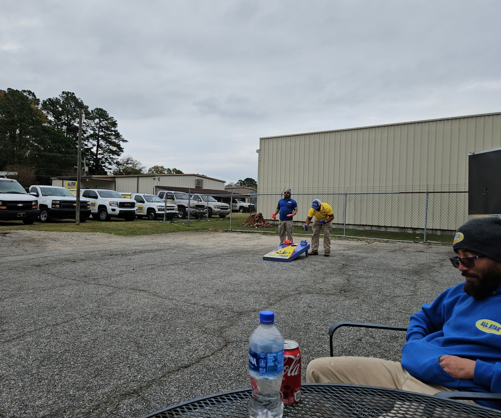 Two people play cornhole on a gravel lot near parked vehicles, while a person in the foreground sits at a table.