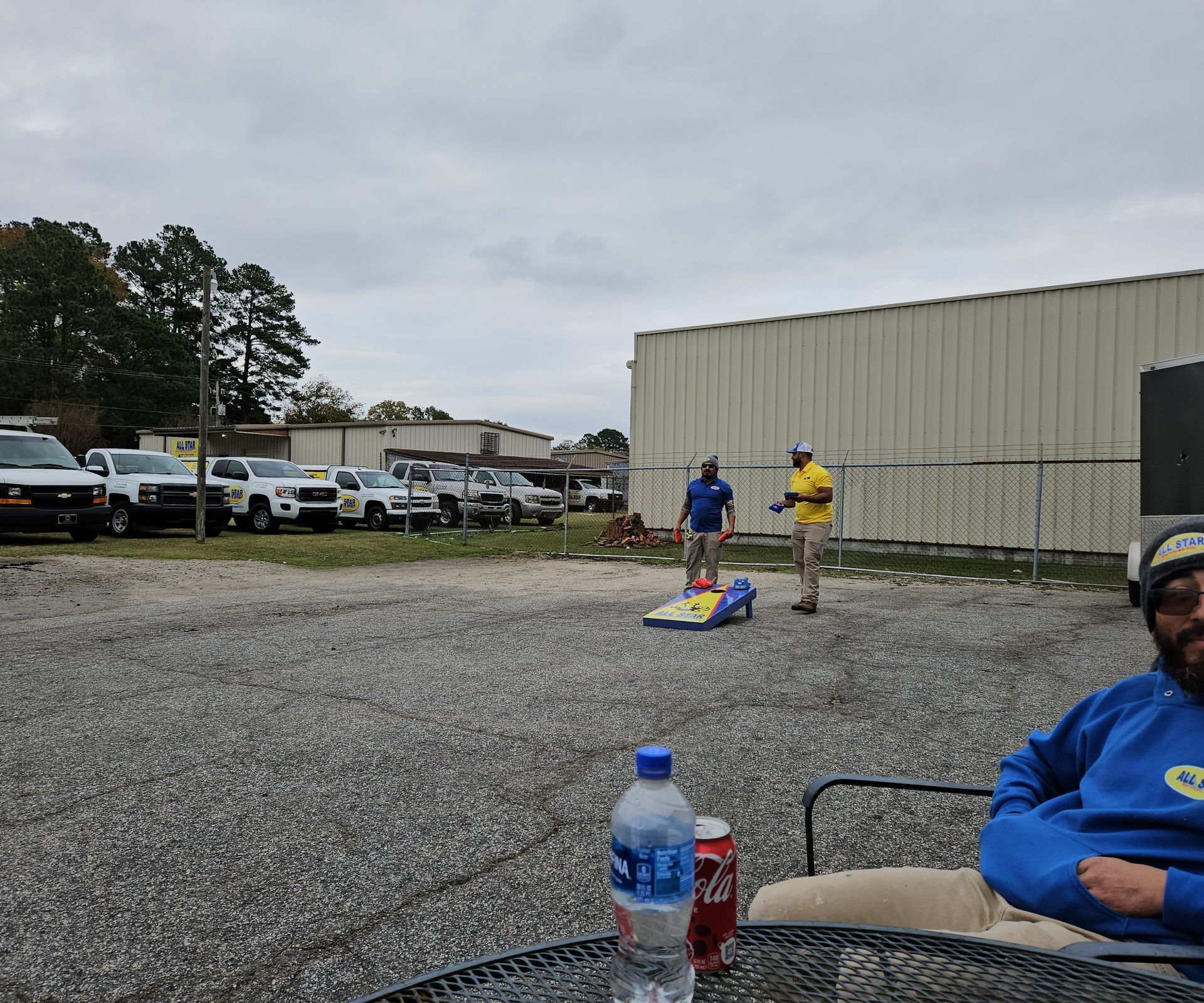 Two people play cornhole on a gravel lot near several parked vehicles, while a person sits in the foreground.