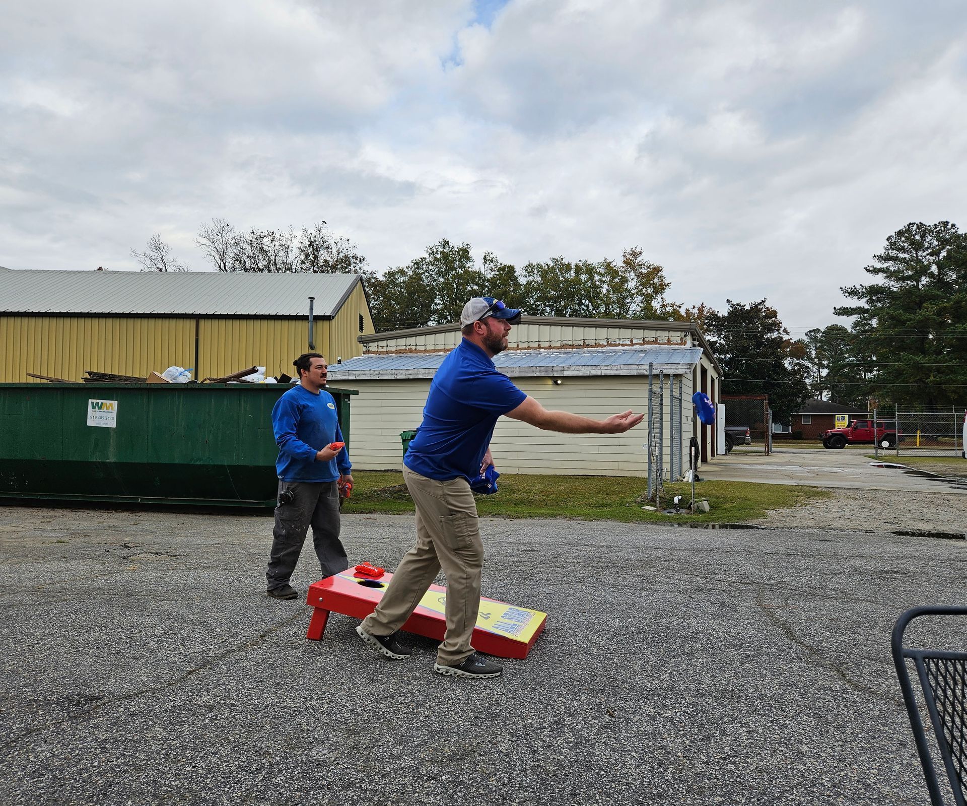 Two people play cornhole on a gravel lot in front of two storage buildings under a cloudy sky.