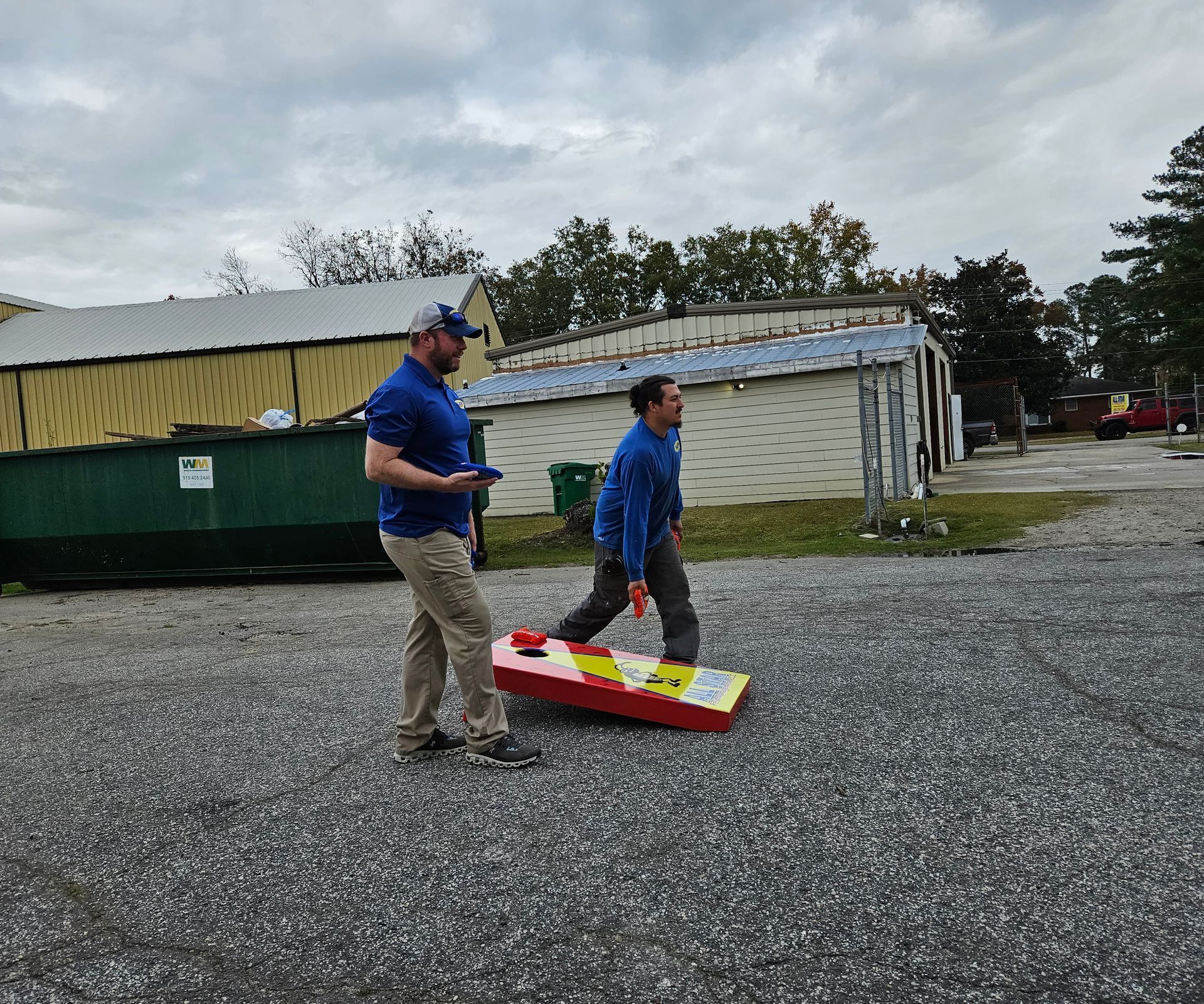 Two people in blue shirts set up a red, yellow, and white cornhole board on a gravel surface outdoors.