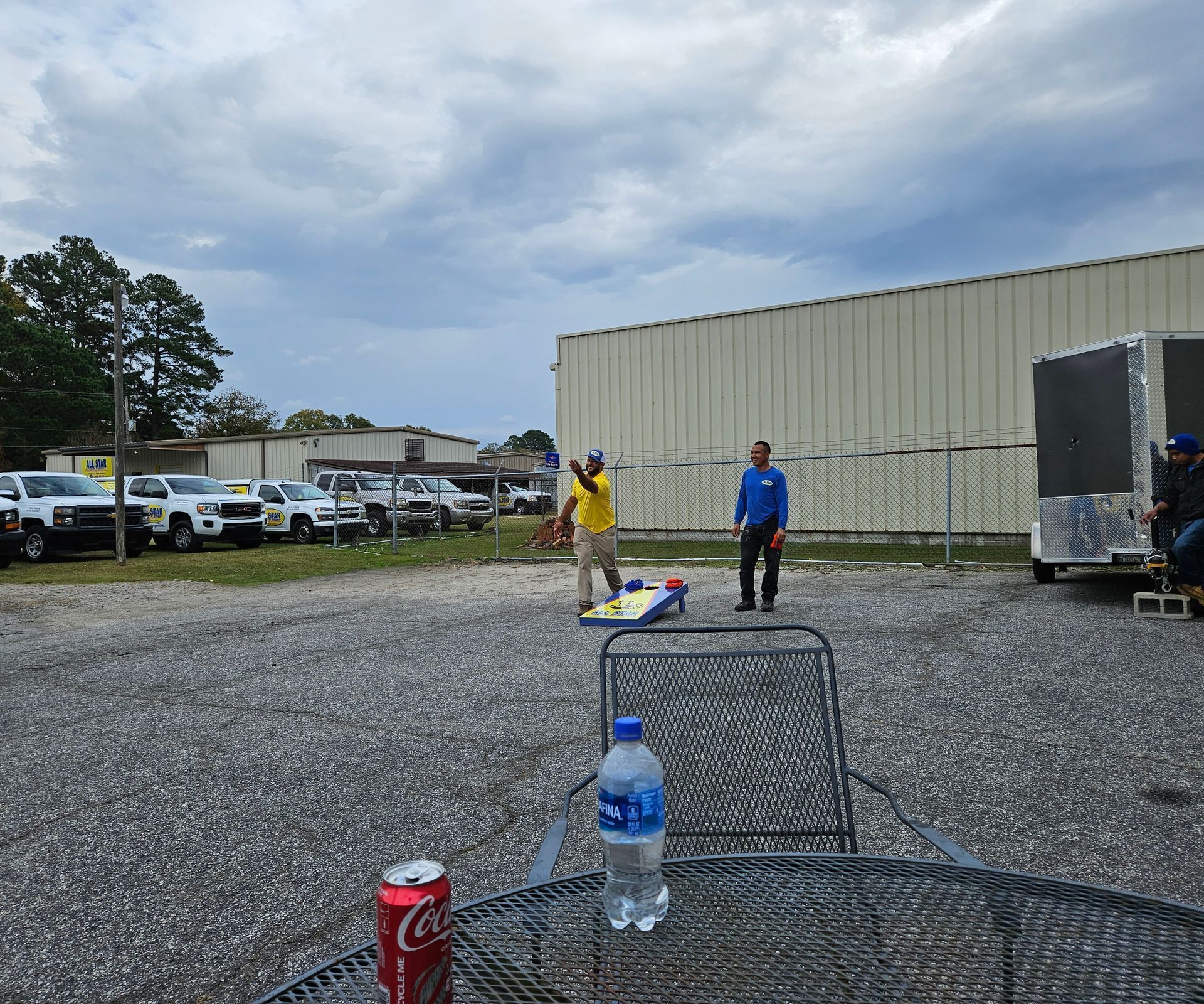 Two people playing cornhole in a gravel parking lot, viewed from a table with a soda can and water bottle.