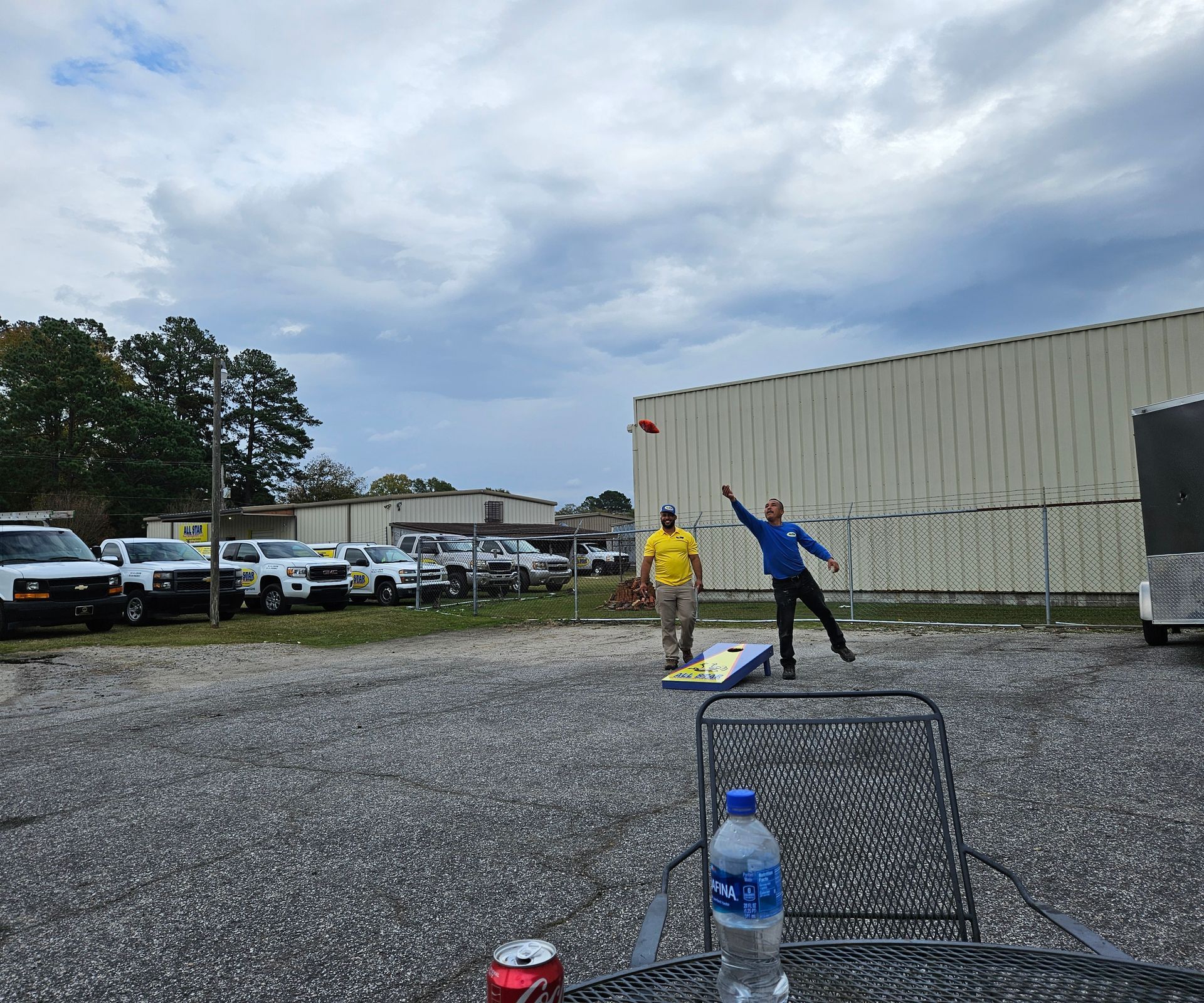 Two people play cornhole on a gravel lot next to a line of trucks and a large metal building under a cloudy sky.