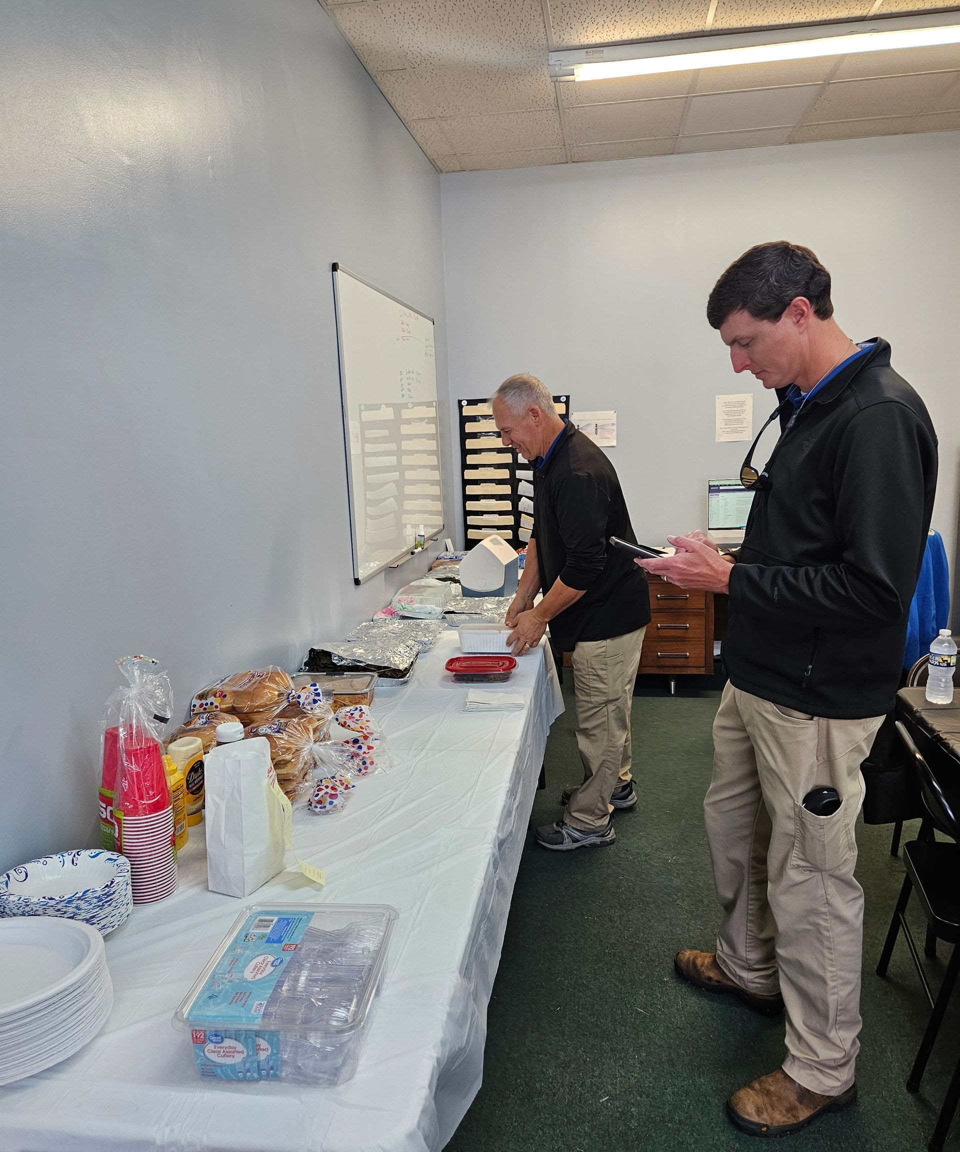 Two people stand by a long table covered with food items and supplies in a room with a whiteboard and light-colored walls.