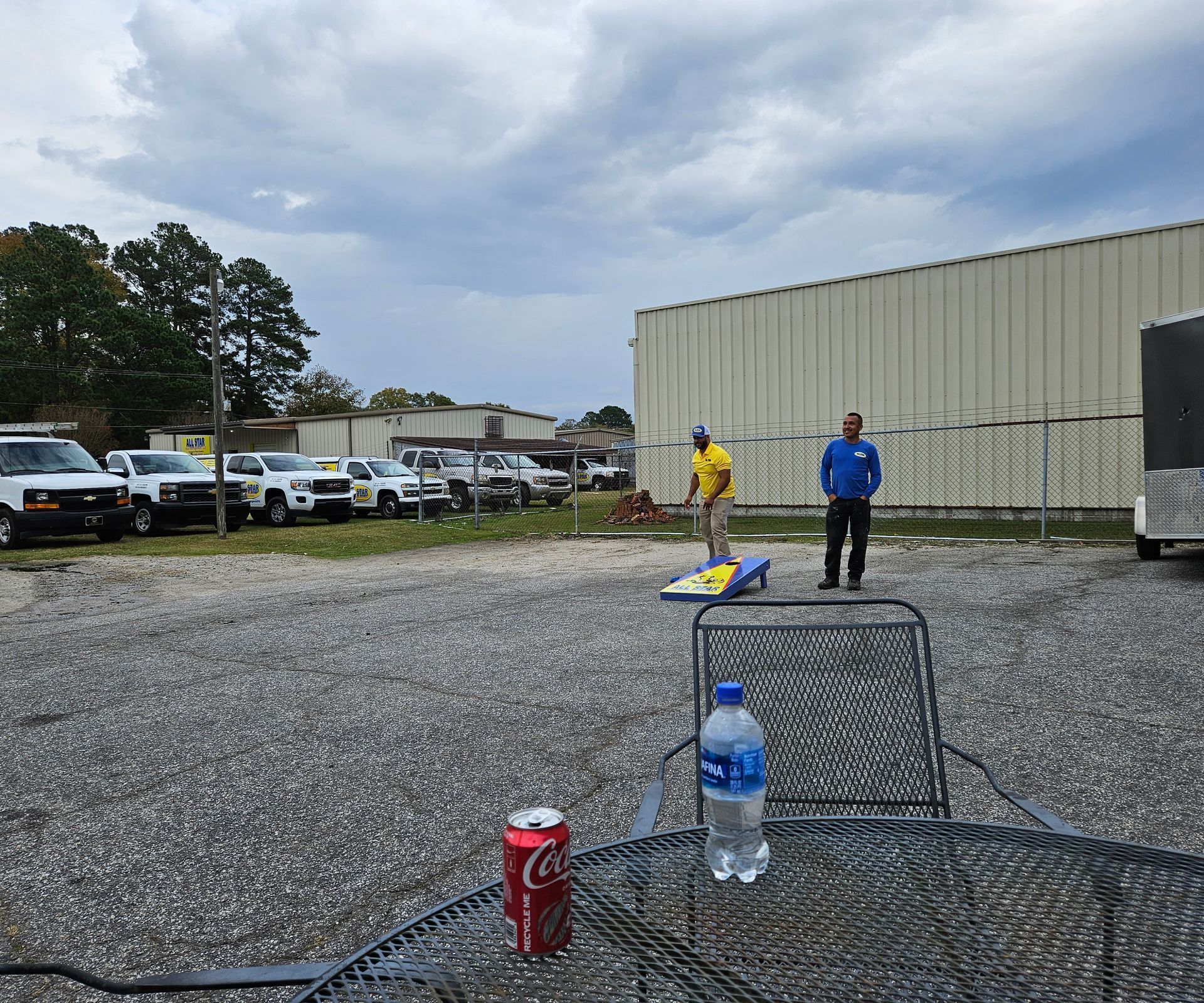 Two people play cornhole on a gravel lot next to a large building, viewed from a table with a soda can and water bottle.