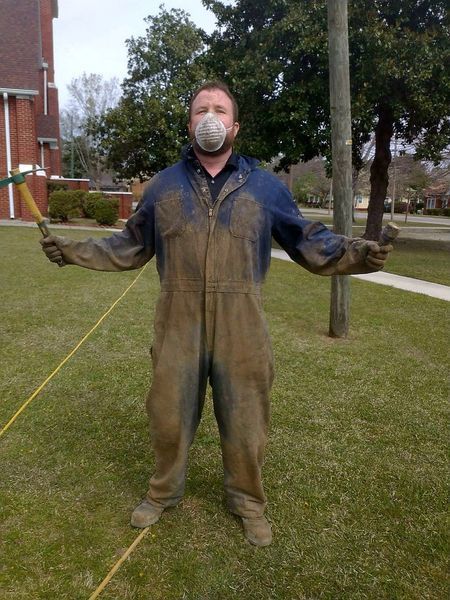 A person in a dust-covered jumpsuit and face mask standing on a lawn holding a small tool in each outstretched hand.