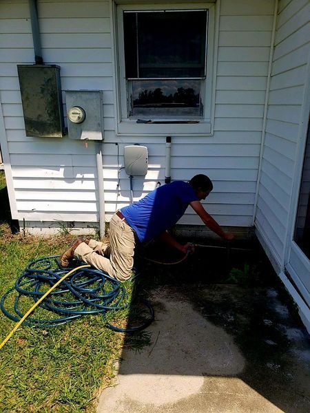 A person in a blue shirt and light pants kneeling on the grass, using a hose to spray near the base of a house wall.