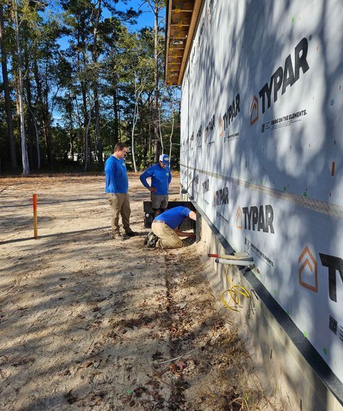 Three construction workers in blue shirts work on the foundation of a building wrapped in white Typar house wrap.