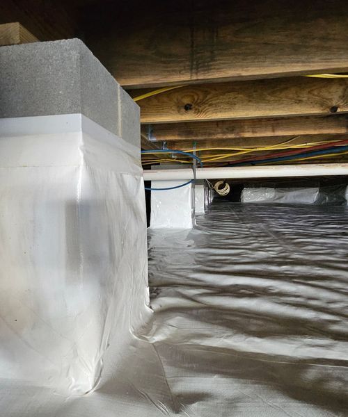 A crawl space with a vapor barrier covering the ground and wrapped around concrete support pillars under floor joists.