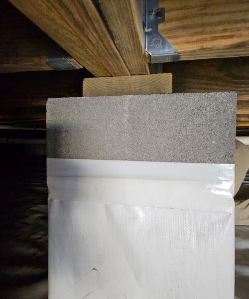 A structural support post with a concrete block base, wooden shim, and joist hanger under a wooden floor frame.
