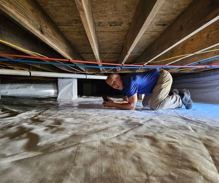 A person in a blue shirt and khaki pants crawls on a white plastic vapor barrier in a crawlspace under wooden floor joists.