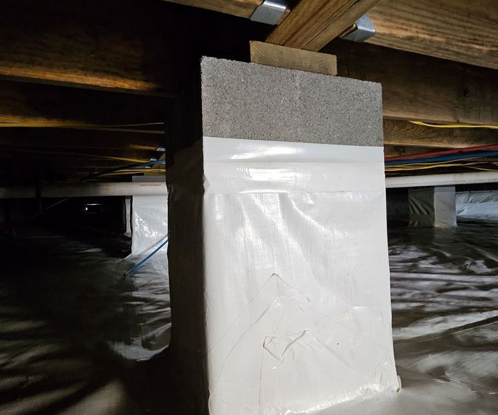 A concrete pier support in a crawl space, wrapped in white vapor barrier material and topped with a wood shim.