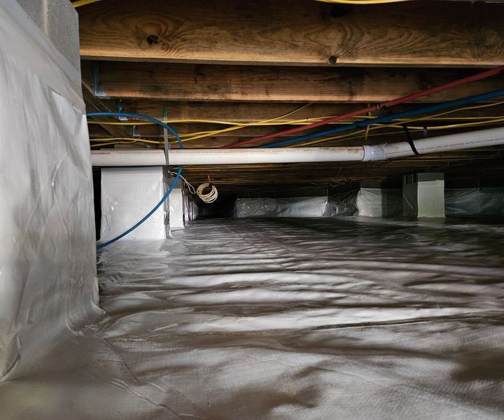 A crawl space featuring a clean white vapor barrier covering the ground, wooden joists, and various utility pipes and wires.