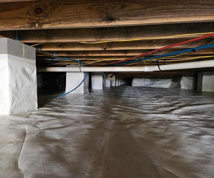 A crawl space under a house with a white vapor barrier on the floor and piers, wooden floor joists, and exposed pipes.