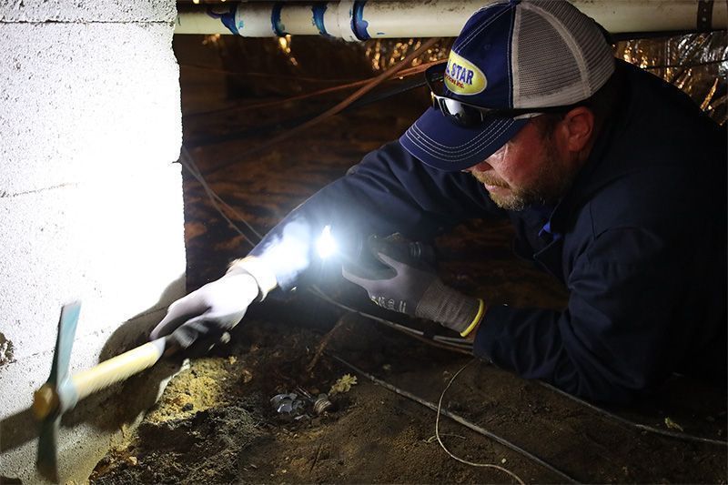 A worker in a cap and gloves uses a tool to inspect the foundation in a dimly lit crawl space.