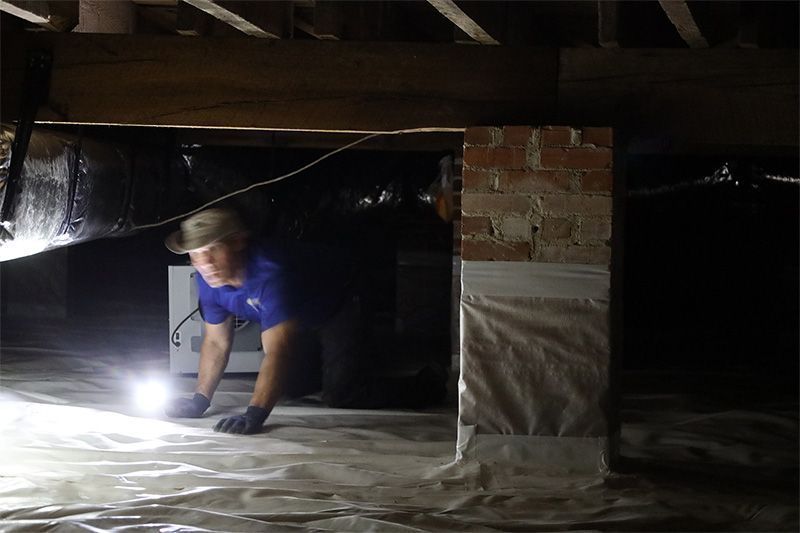 A technician with a flashlight kneels in a crawl space next to a brick support pier and a white HVAC unit.