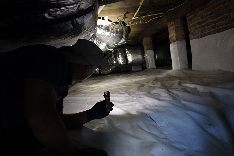 A person in a crawl space inspects a white vapor barrier with a flashlight.