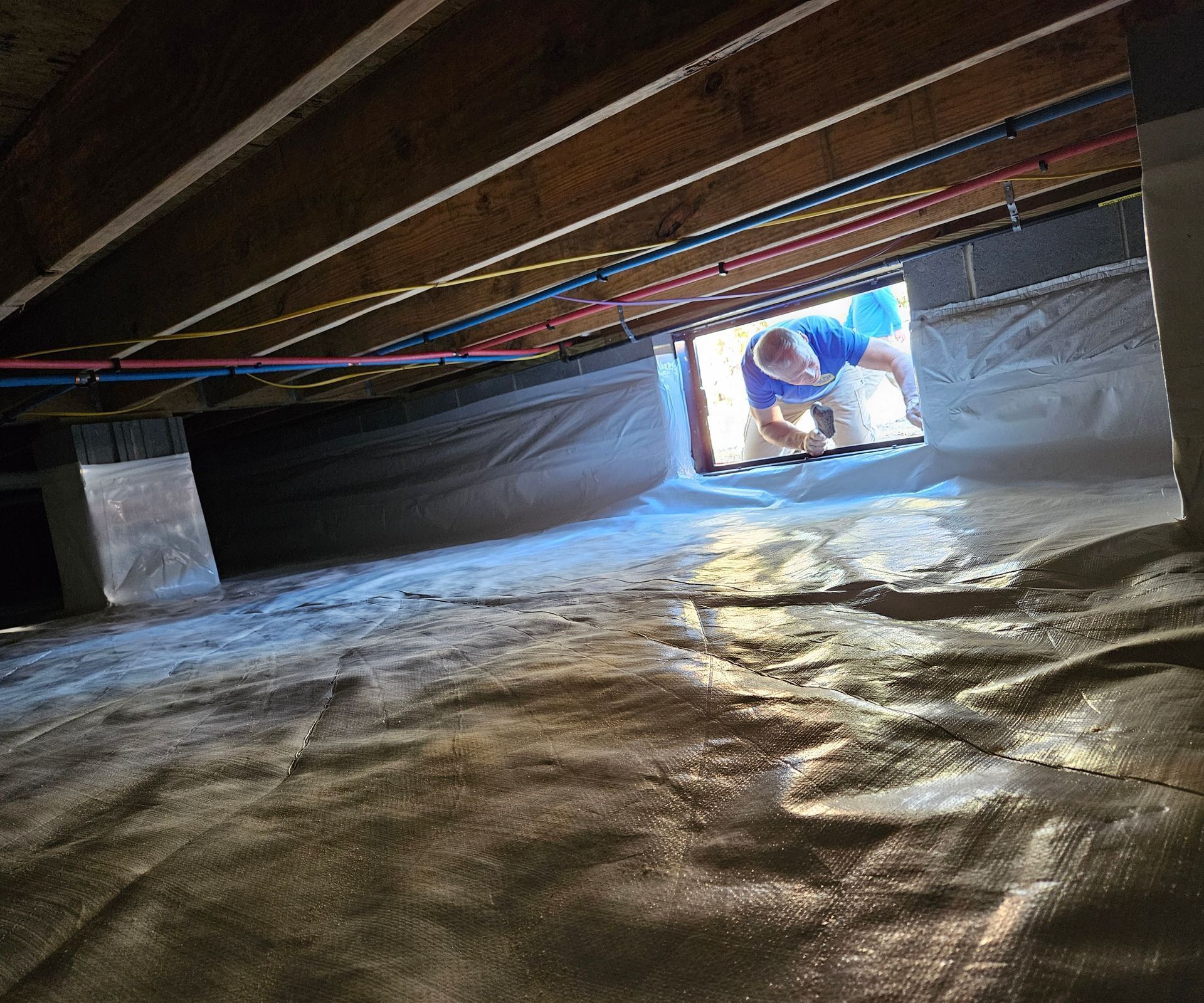A person looks into a crawl space featuring a white vapor barrier covering the ground and walls beneath wooden joists.