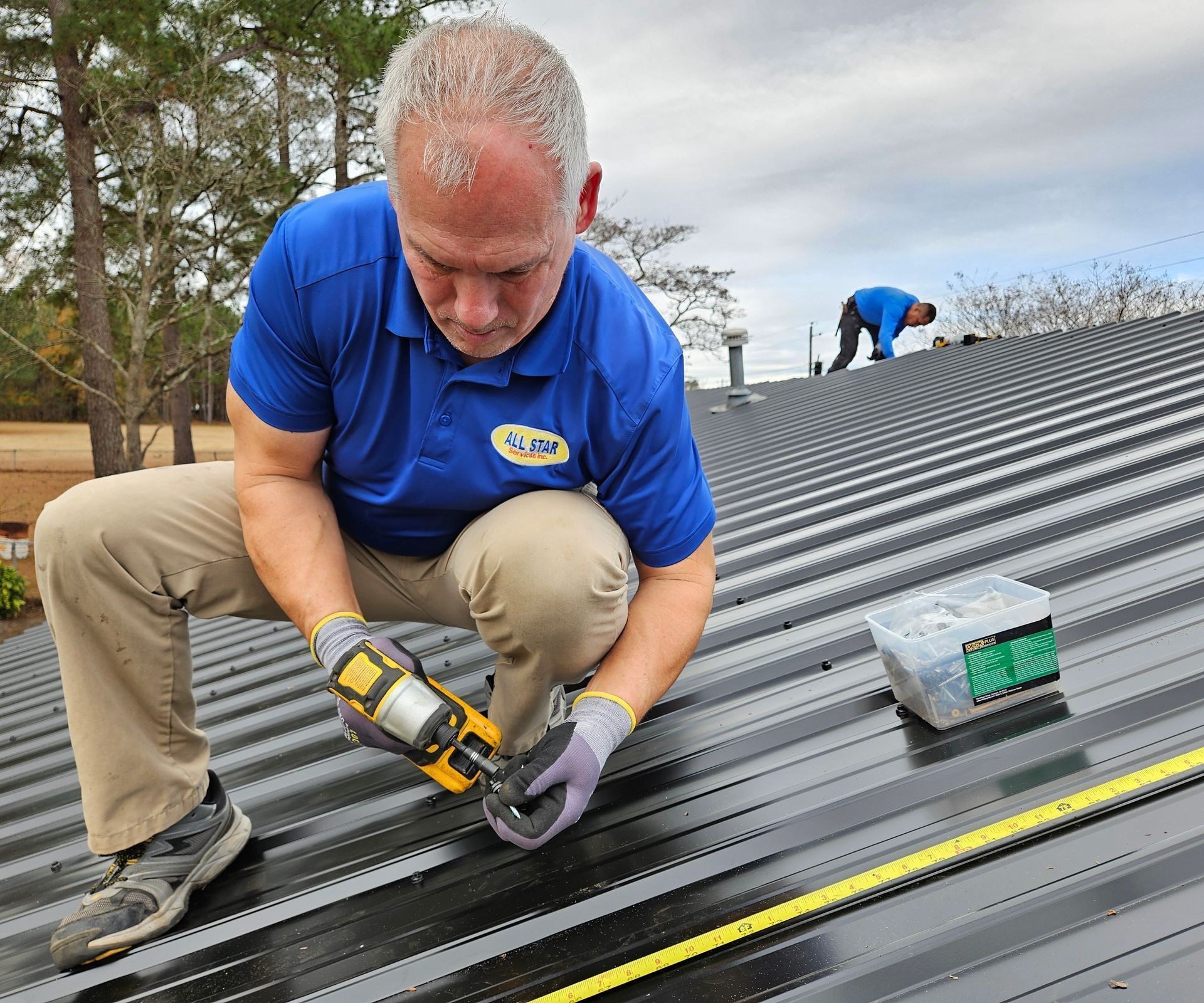 A worker in a blue polo shirt uses a power tool to fasten a black metal roof, with a second person working nearby.
