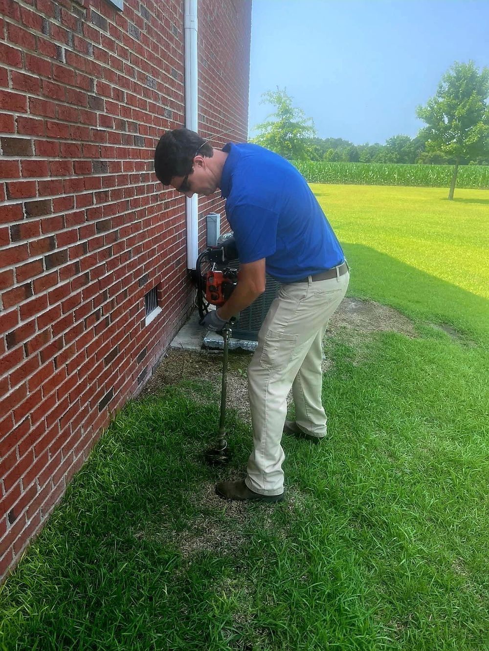 A technician in a blue polo and tan pants uses a gas-powered soil auger to drill into the ground next to a brick building.