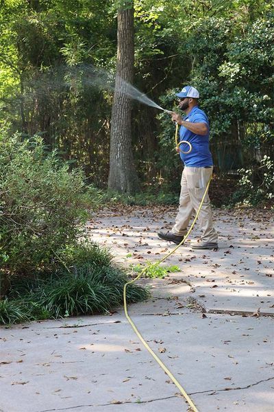 A person in a blue shirt and tan pants sprays water with a hose onto plants along a paved driveway.
