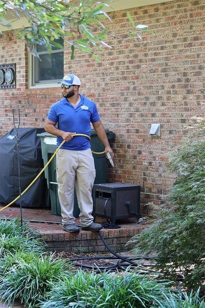 A worker in a blue polo shirt and khaki pants stands outside a brick house, holding a yellow hose.