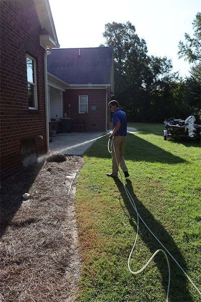 A person in a blue shirt walks across a grassy yard next to a brick house, holding a hose connected to the foundation.