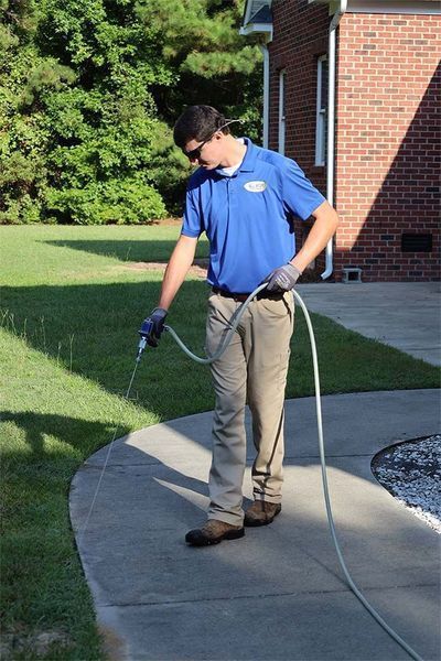 A professional in a blue uniform sprays a solution onto a concrete walkway near a brick building.