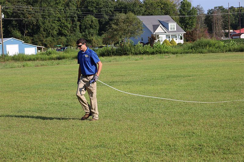 A person in a blue shirt and khaki pants walks across a grassy field while trailing a white rope attached to their waist.