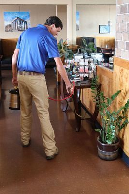 A worker in a blue polo and tan pants uses a spray hose to apply pest control treatment inside a restaurant.