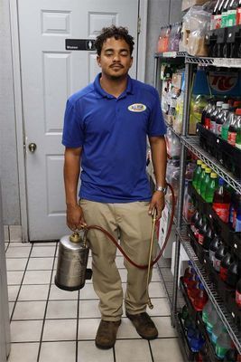 A technician in a blue polo and tan pants holds a metal pest control sprayer in a store storage area beside metal shelves.